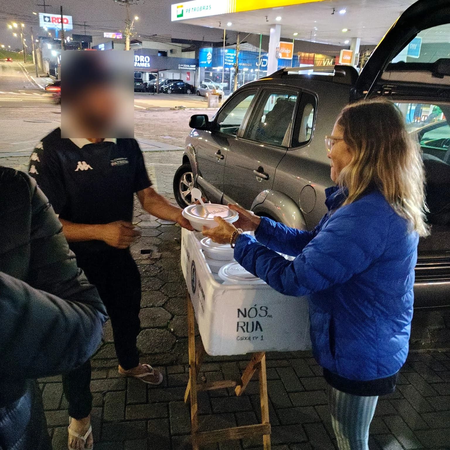 Voluntária entregando marmita ao lado da caixa térmica com a identidade do Nós na Rua.